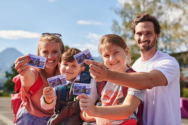 Eine Familie – Mama, Papa und ihre zwei Kinder – lachen fröhlich und halten ihre Erlebnis Cards für die Region Villach in den Händen. 